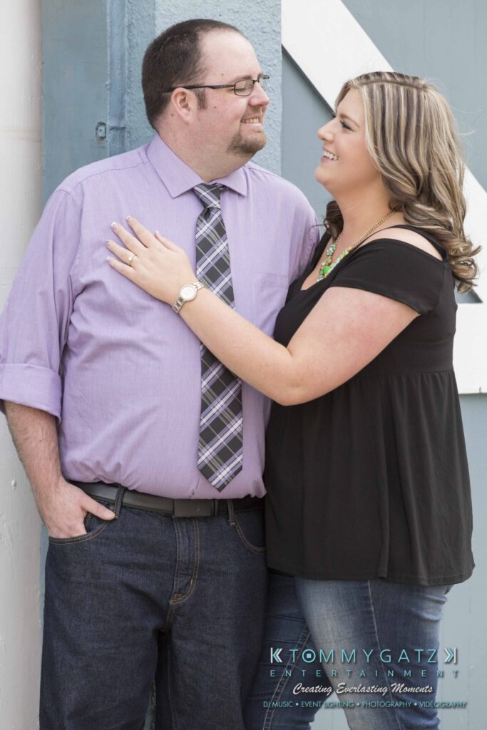 A man and woman stand closely, smiling at each other. The woman has her hand on the man's chest. Both are casually dressed.