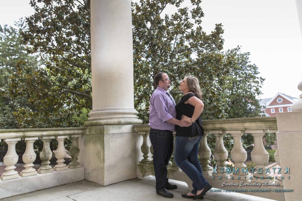 A couple stands on an outdoor balcony in front of a large tree, facing and embracing each other.