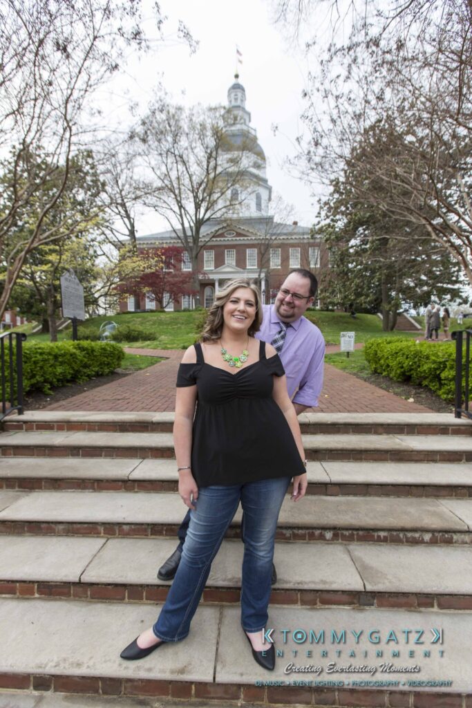 A smiling woman and man pose on stone steps, with a historic building and trees in the background.