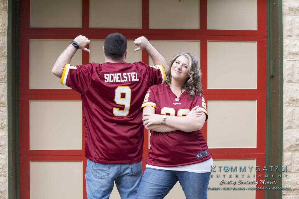Two people wearing matching red sports jerseys stand in front of a brown garage door. The man shows the name "Sichelstiel" on his back, while the woman stands with crossed arms, smiling.