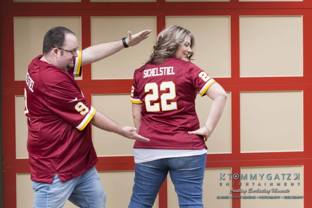 Two people in matching red sports jerseys with names and numbers on the back stand in front of a red and yellow background. The man gestures toward the woman.