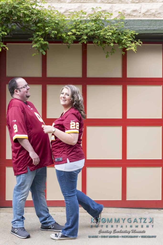 A man and woman wearing matching maroon sports jerseys stand in front of a red and beige garage door, smiling at each other.