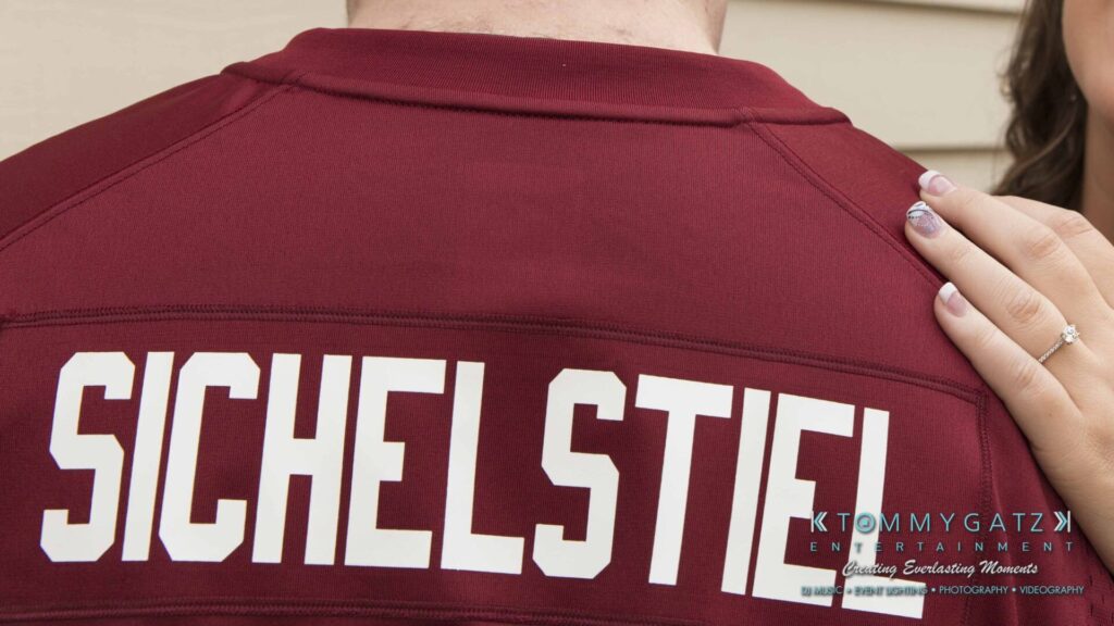 Back of a person wearing a maroon shirt with "Sichelstiel" printed on it, next to a hand resting on their shoulder.