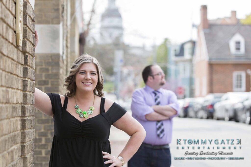 Woman in black dress leans against a brick wall, smiling, while a man in a purple shirt stands in the background with his arms crossed.