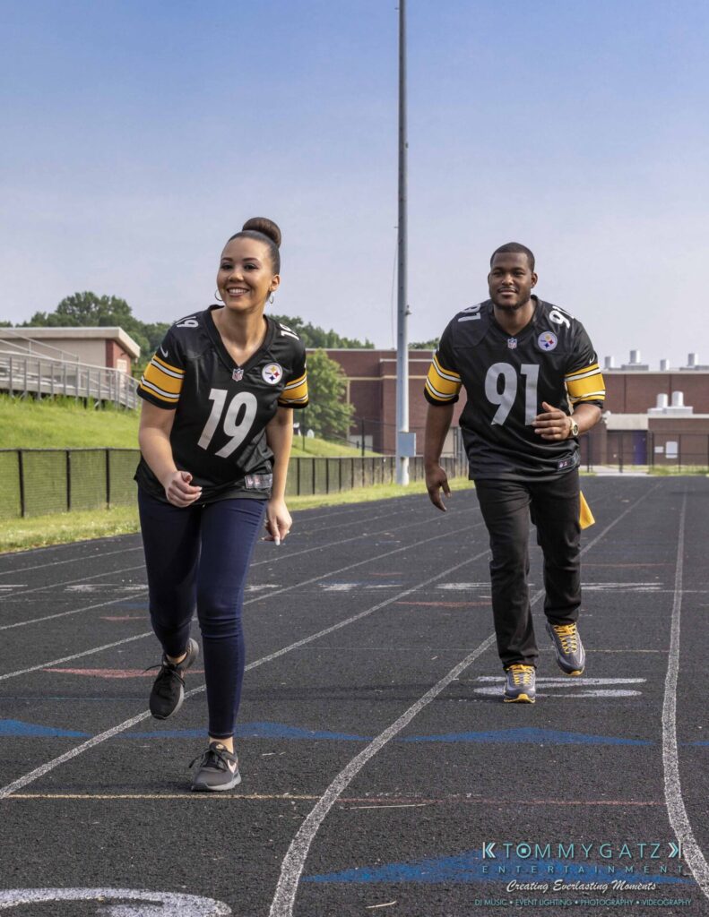 Two people wearing matching sports jerseys run on a track outdoors.