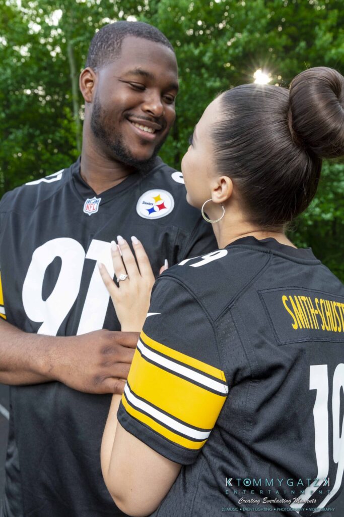 A couple dressed in matching football jerseys smiles at each other while standing outdoors.