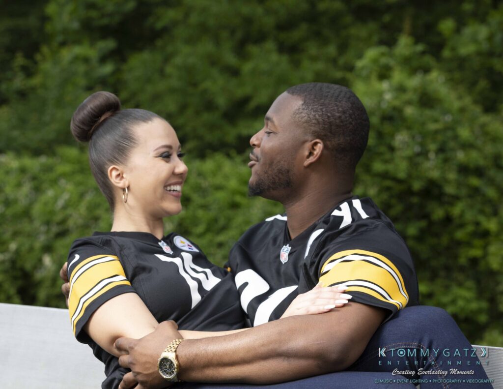 A couple wearing matching sports jerseys sits closely on a bench outdoors, smiling at each other against a lush green background.