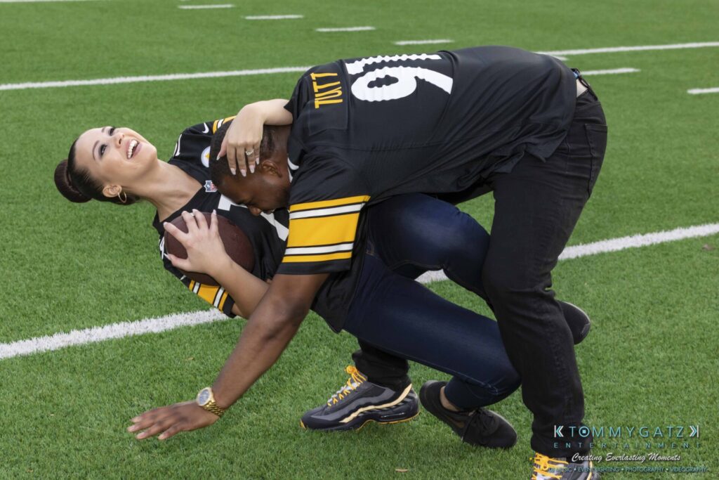 Two people wearing football jerseys playfully interact on a football field. One holds a football while the other bends slightly, both smiling.