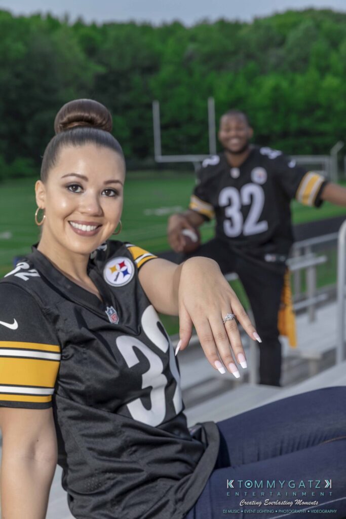 A woman shows her engagement ring while sitting in a stadium, with a man holding a football in the background. Both are wearing football jerseys with the number 32.