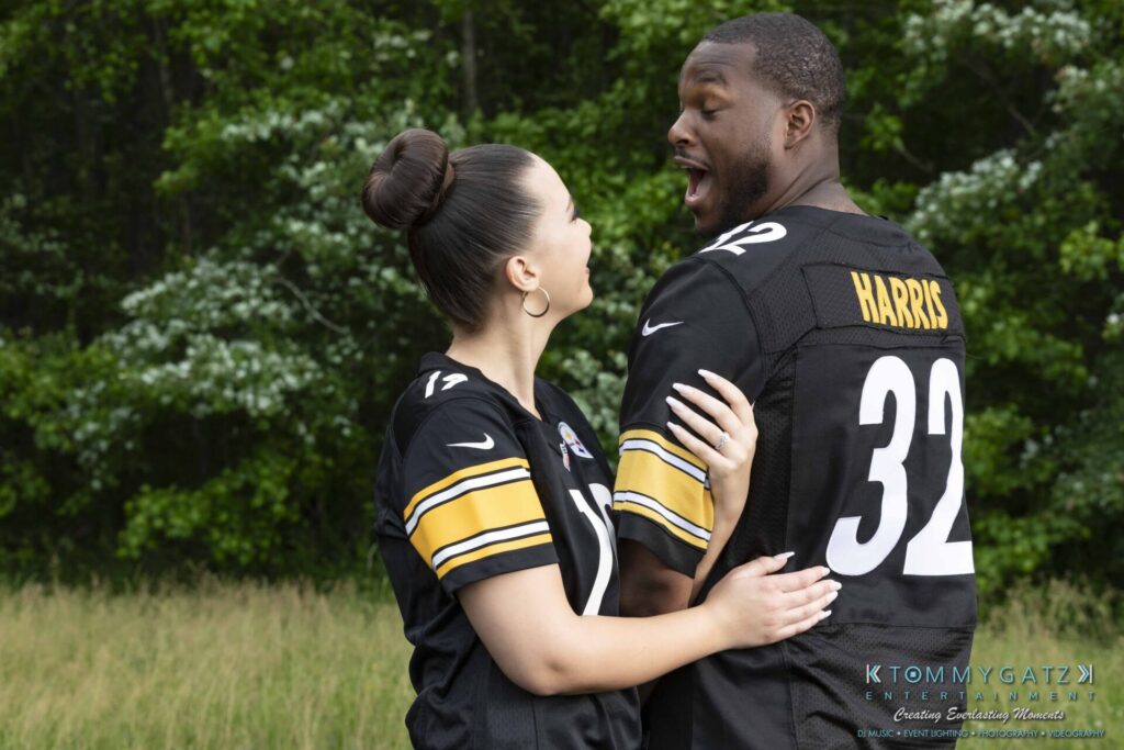 Two people wearing matching black sports jerseys stand in a field, facing each other. One person appears surprised or excited, while the other is smiling and holding their arm.
