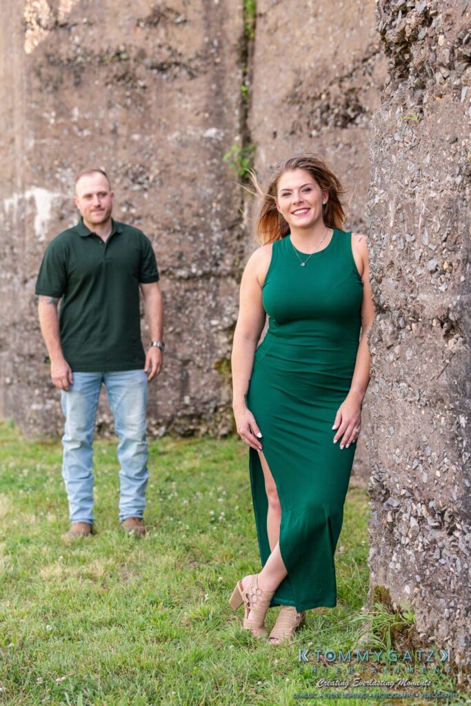A woman in a green dress leans against a stone wall outdoors, with a man in a green shirt and jeans standing in the background on the grass.