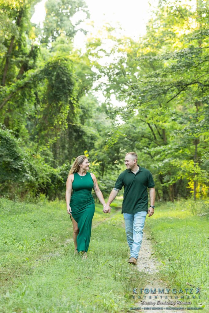 A man and woman hold hands walking on a grassy path in a wooded area. Both are dressed in green outfits, with the woman in a long dress and the man in a polo shirt and jeans.