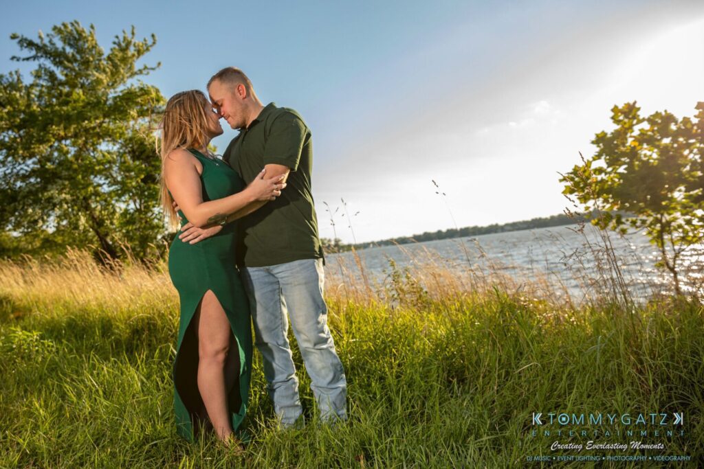 A couple embraces on a grassy lakeside under a partly cloudy sky. The woman wears a green dress, and the man wears a green shirt and jeans. Trees and water are visible in the background.