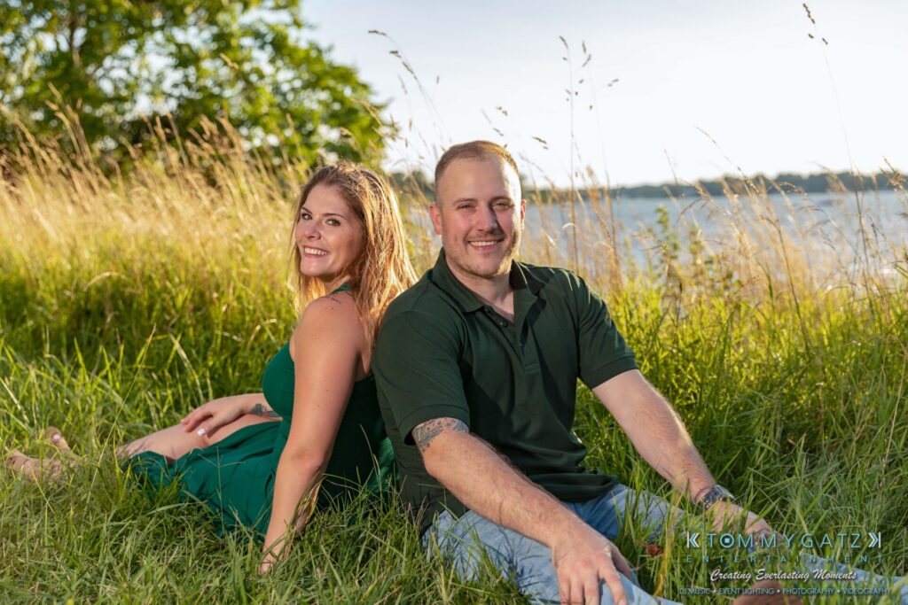 A man and a woman sit on the grass near a body of water, smiling and facing the camera.