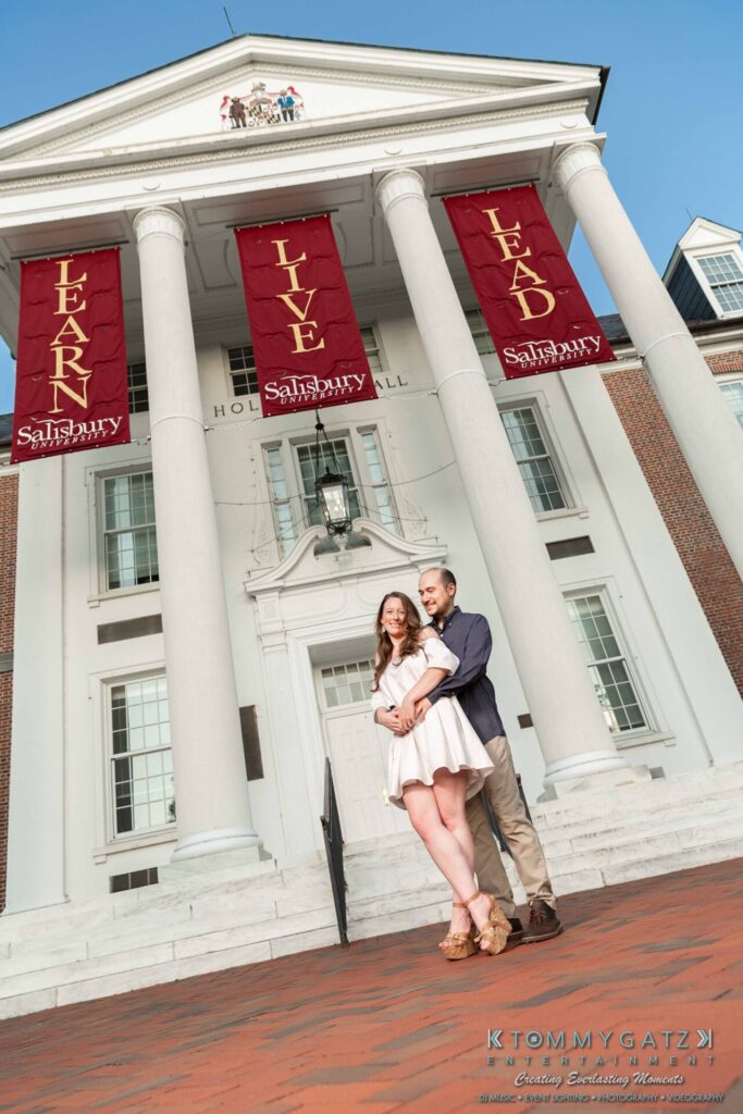 A couple stands together in front of a brick building with tall columns and red banners reading "Learn," "Live," "Lead.