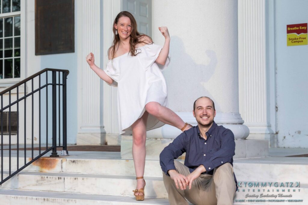 A woman in a white dress poses confidently with her arms flexed, while a seated man in a dark shirt smiles against a white column backdrop.