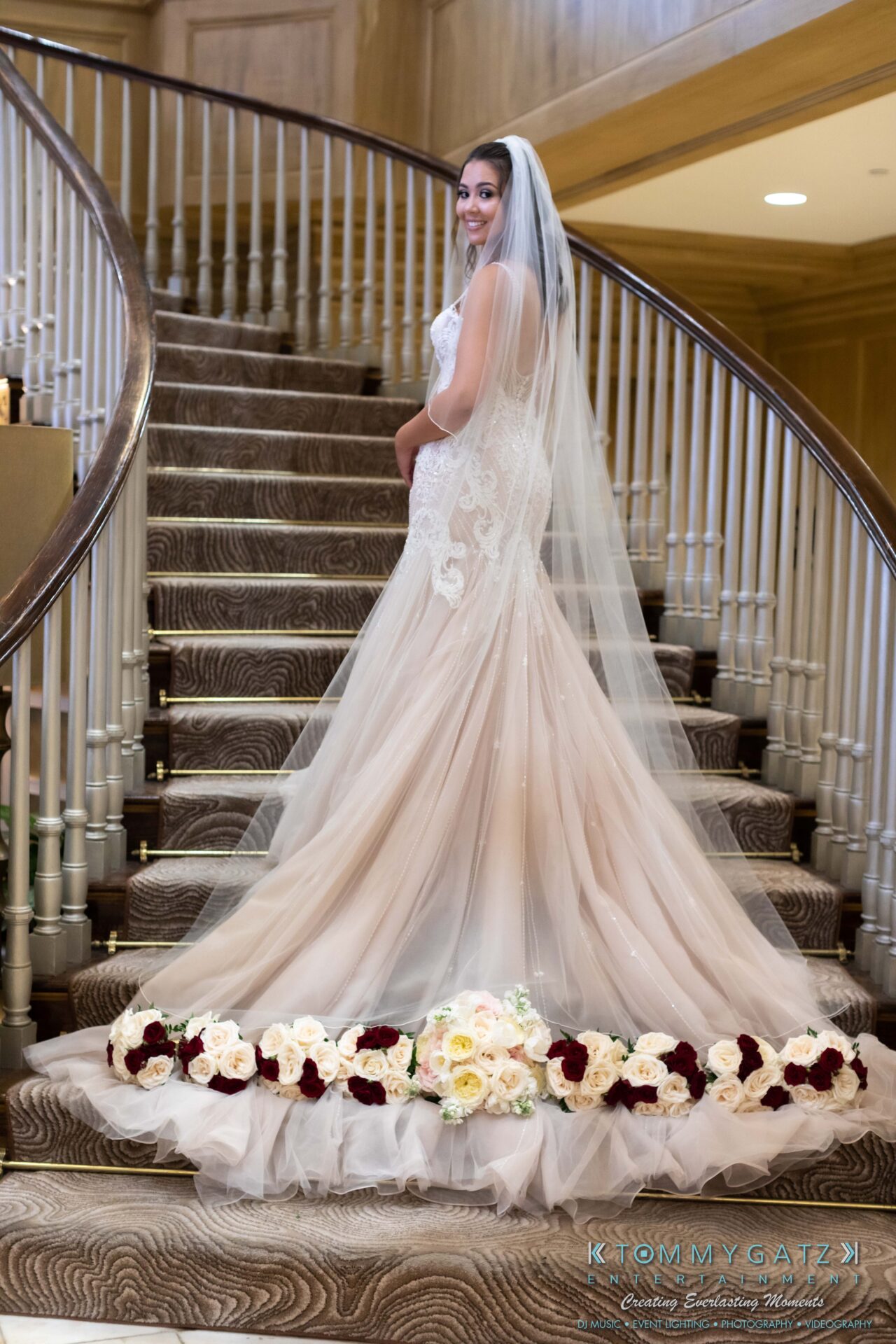 Bride poses on a staircase in a lace wedding gown with a long train and veil, surrounded by floral arrangements.