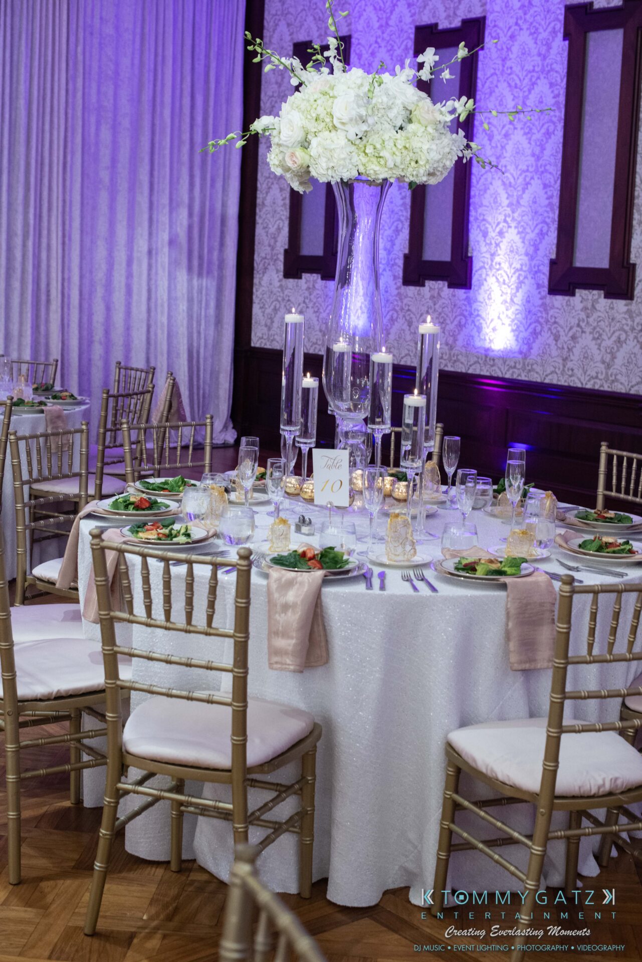 A round elegantly set dining table with a tall floral centerpiece, surrounded by gold chairs. Plates contain salads and there are wine glasses and votive candles on the table.