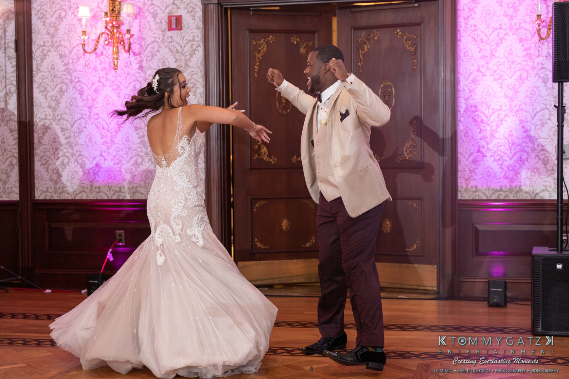 A couple dances energetically in a formal setting, with the woman in a white dress and the man in a beige jacket.