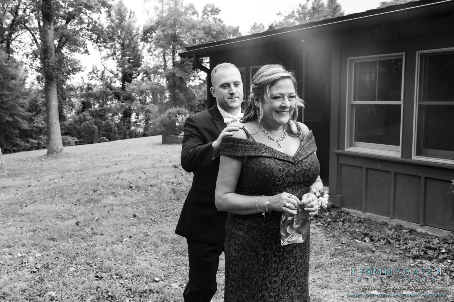 A man in a suit pins a corsage on a smiling woman in a dark dress outside a house, with trees in the background.