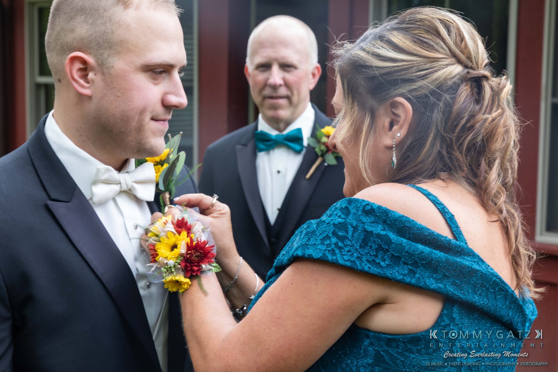 A woman adjusts the boutonniere of a man in a tuxedo, while another man stands in the background.