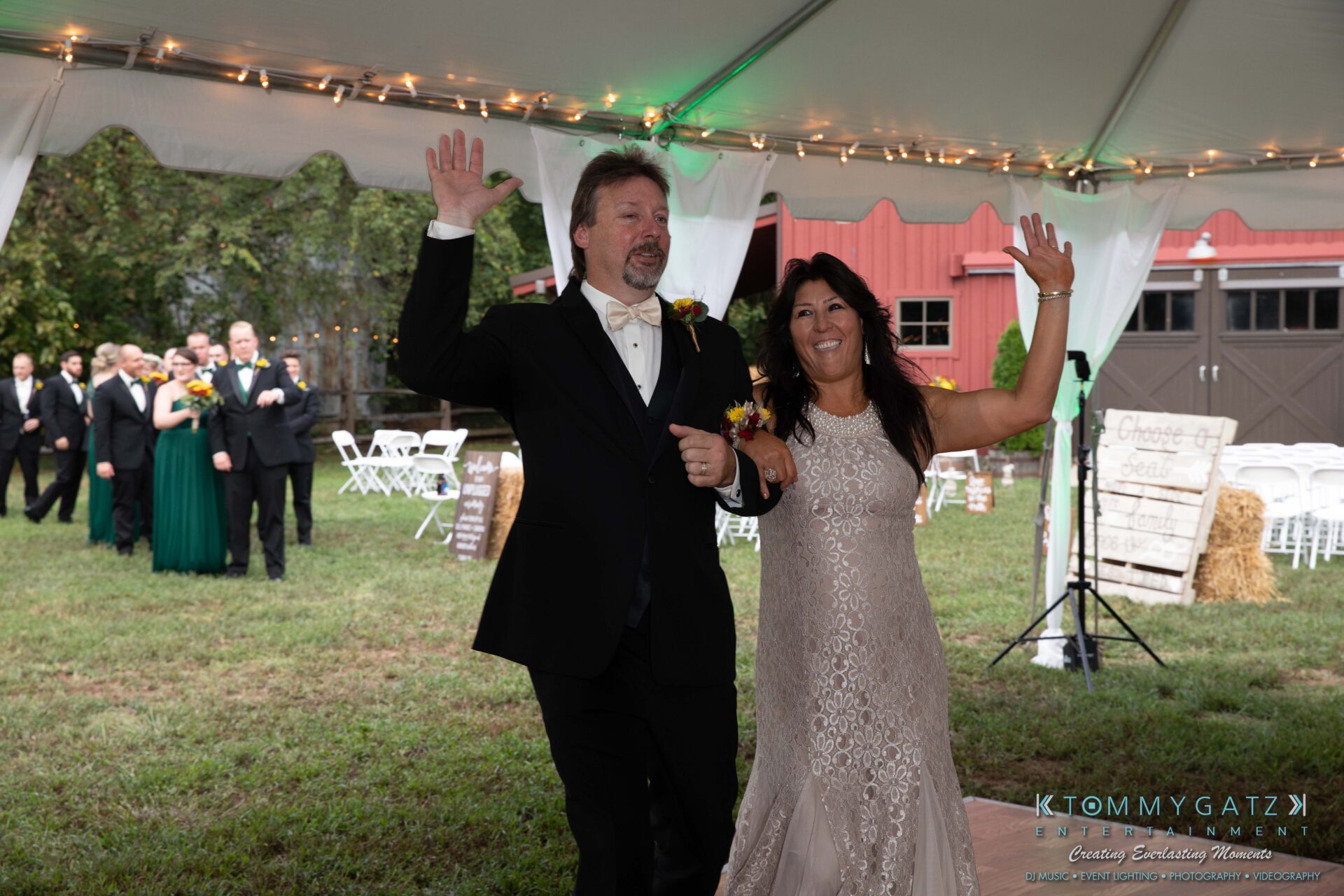A couple in formal attire enters an outdoor event tent, smiling and waving, with guests and greenery in the background.