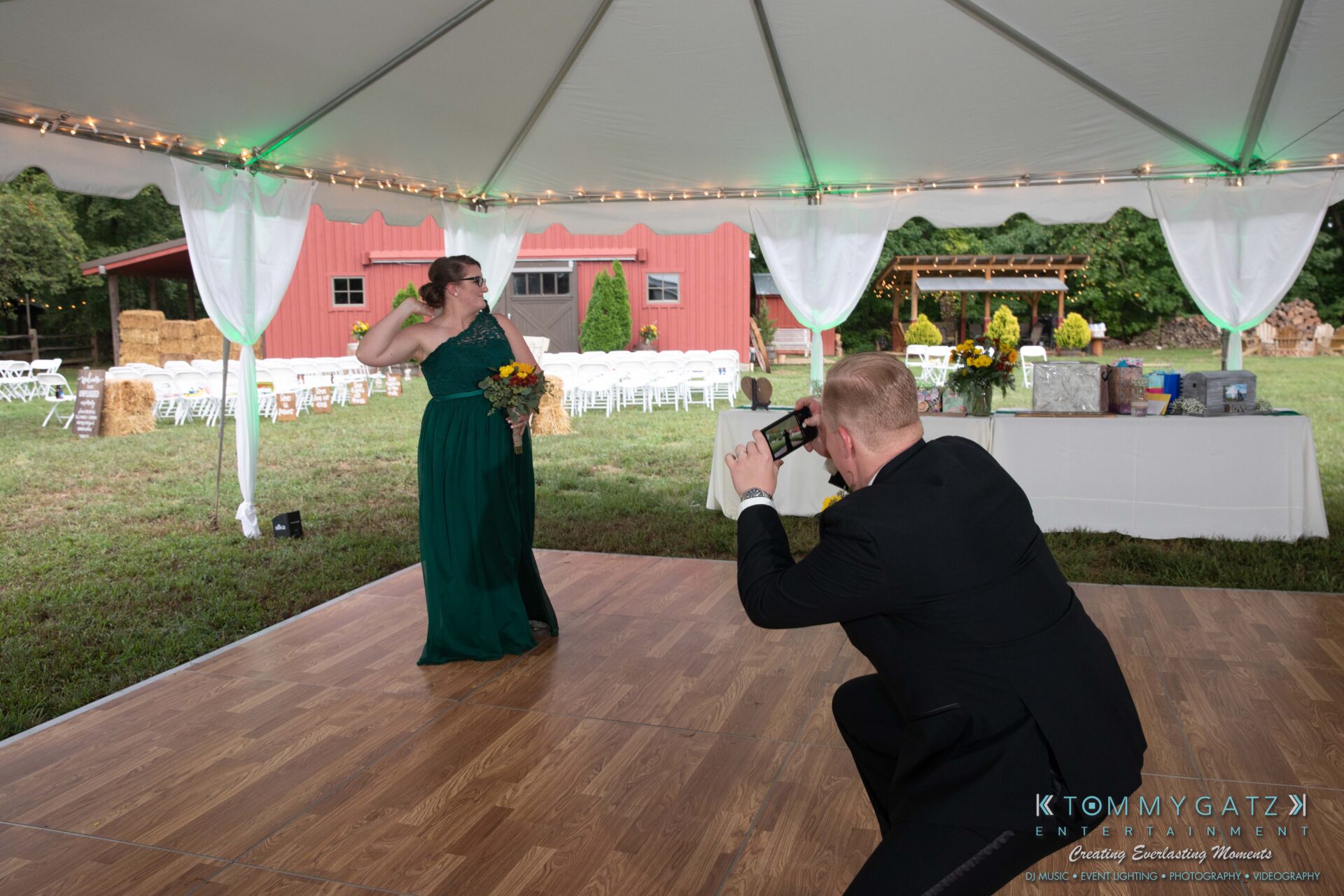 A woman in a green dress poses with a bouquet while a man in a suit photographs her under a tent at a rustic outdoor event.