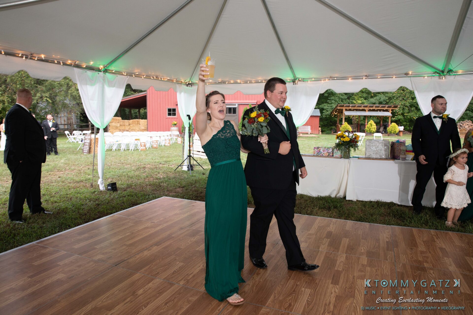 A woman in a green dress raises a glass while holding a bouquet next to a man in a suit under a tent at an outdoor event.