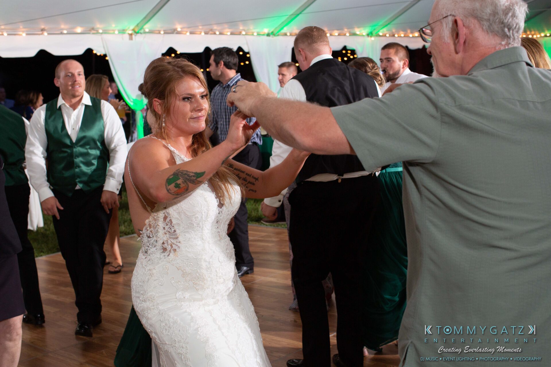 A bride in a white dress dances with an older man under a tent. Guests in formal attire stand and watch.