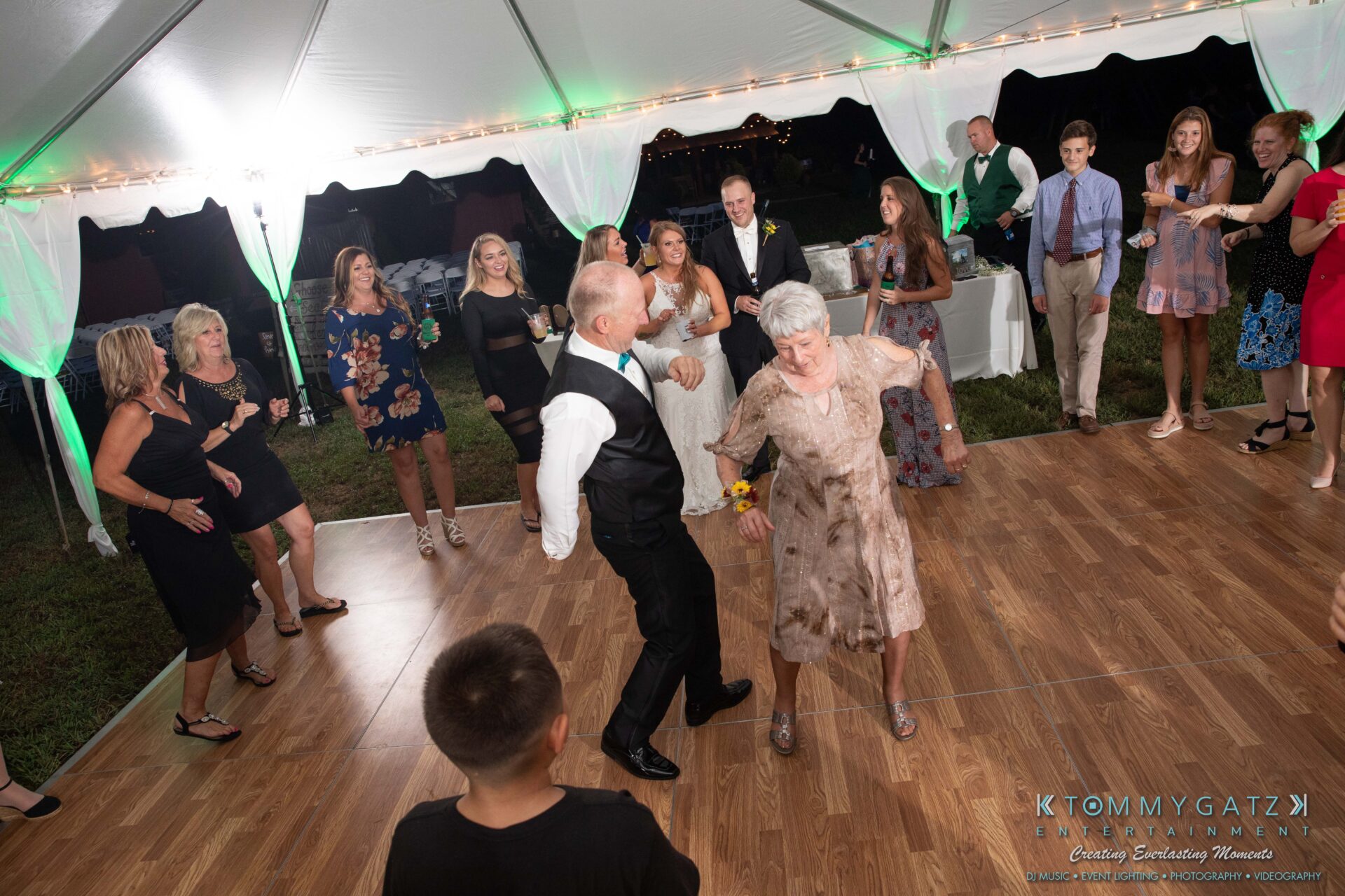People dancing on a wooden floor under a white tent at an outdoor event. A woman in a floral dress and a man in a vest are leading the dance in the foreground.