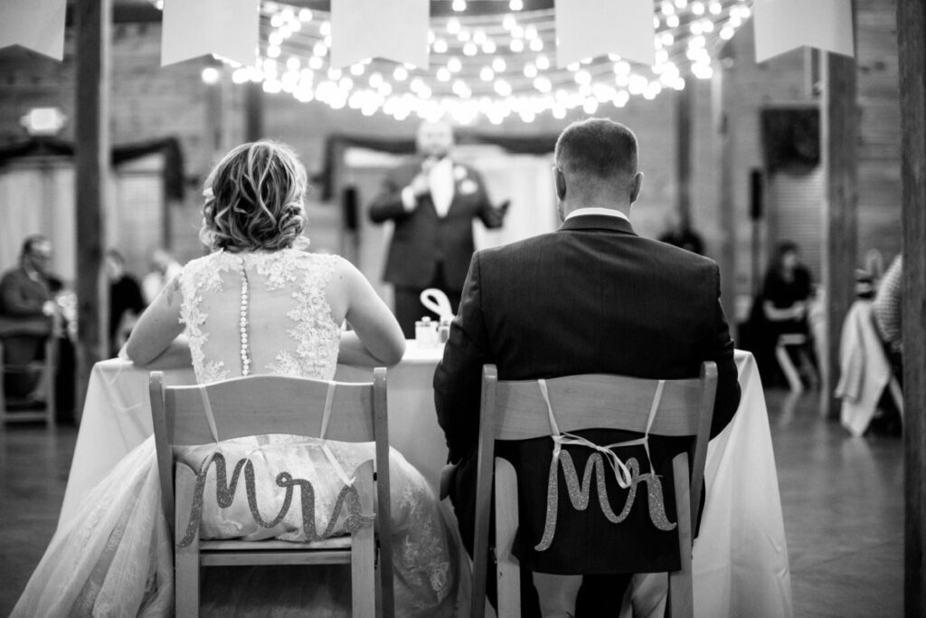 A bride and groom sit at a table labeled "Mr." and "Mrs." facing a speaker. String lights hang above in a decorated venue.