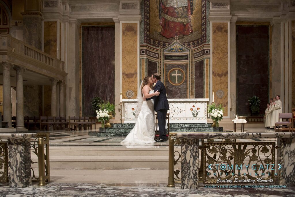 A bride and groom share a kiss at the altar in a grand church setting, with the priest and a witness in the background.