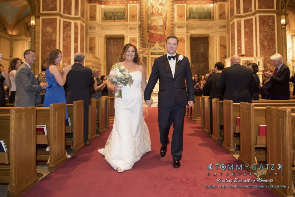 A bride and groom holding hands walk down the aisle of a church, surrounded by guests seated in pews.