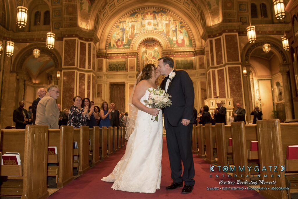 A bride and groom kiss at the altar in a church decorated with murals and arches, while guests watch from the pews.