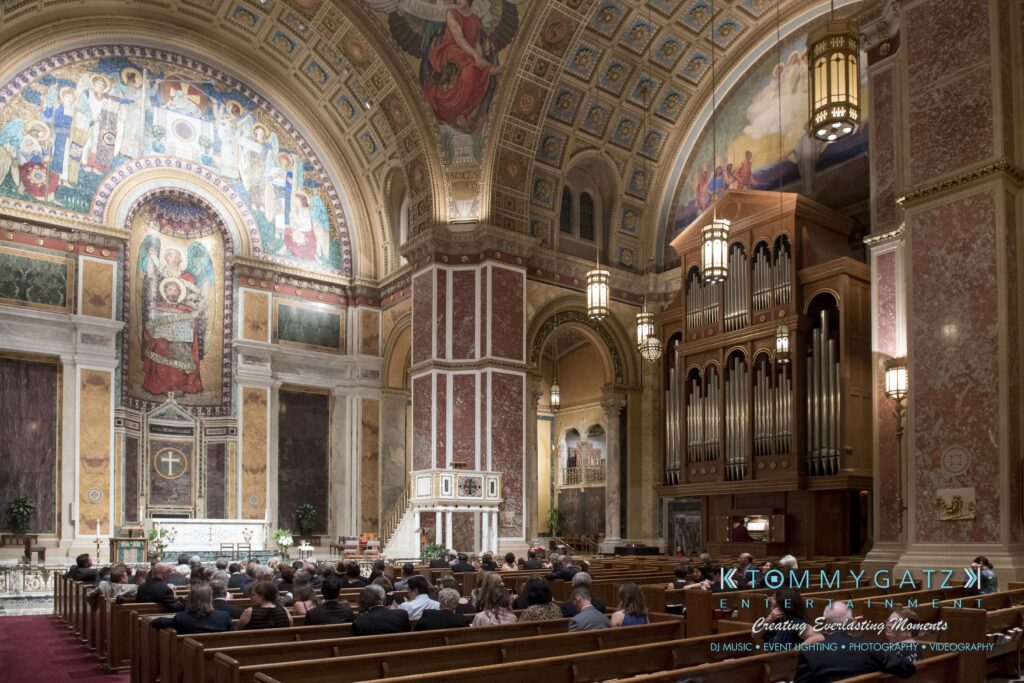 Interior of a grand church with ornate architecture, stained glass windows, and an organ. A congregation is seated, facing the altar under a vaulted ceiling.
