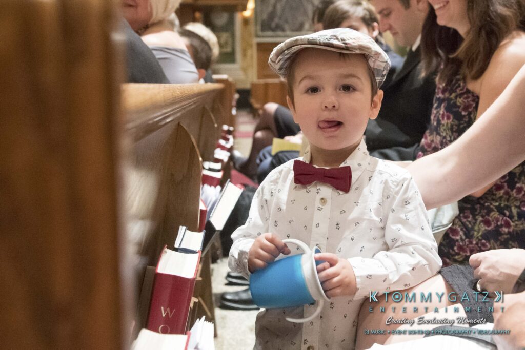 A young boy in a polka dot shirt and bow tie, wearing a flat cap, stands in a church pew holding a blue cup. People are seated around him.