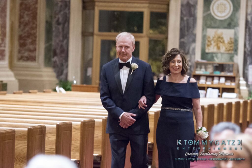 A man in a tuxedo and a woman in an elegant dress walk arm-in-arm down the aisle of a large, ornate church.