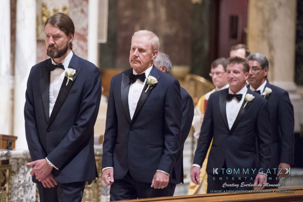 A group of men in formal suits with white rose boutonnieres stand in a church setting.