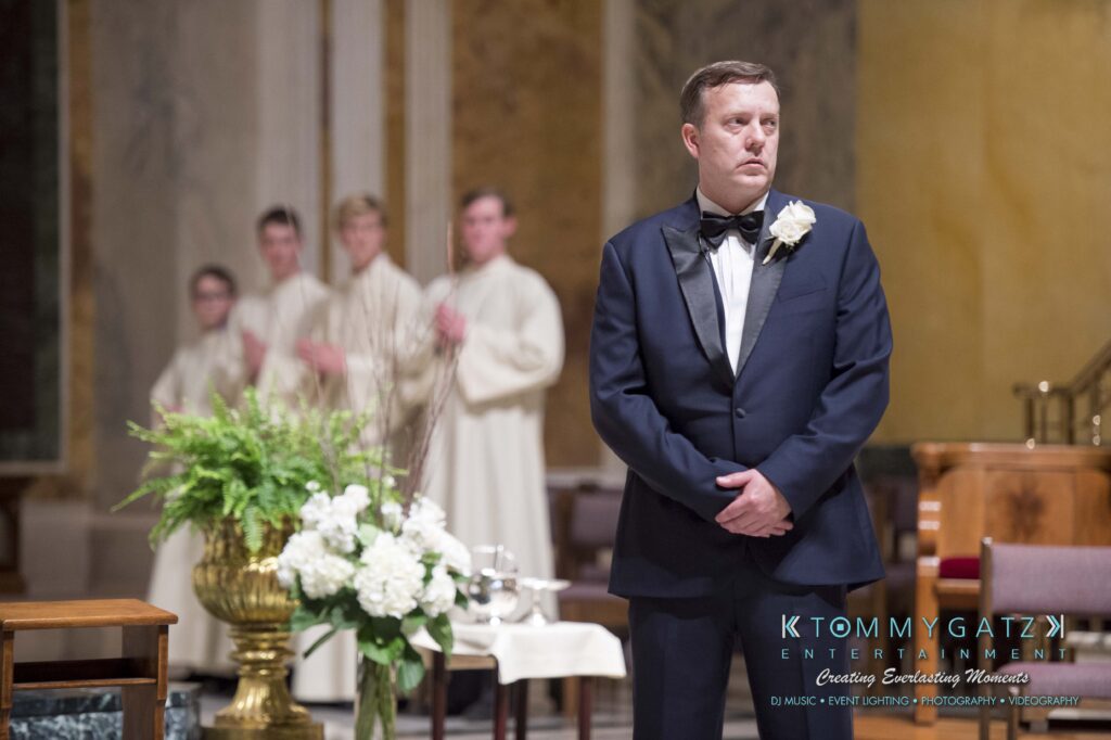 A man in a formal suit stands in a church setting, looking slightly to the side. People in white robes are blurred in the background near a floral arrangement.