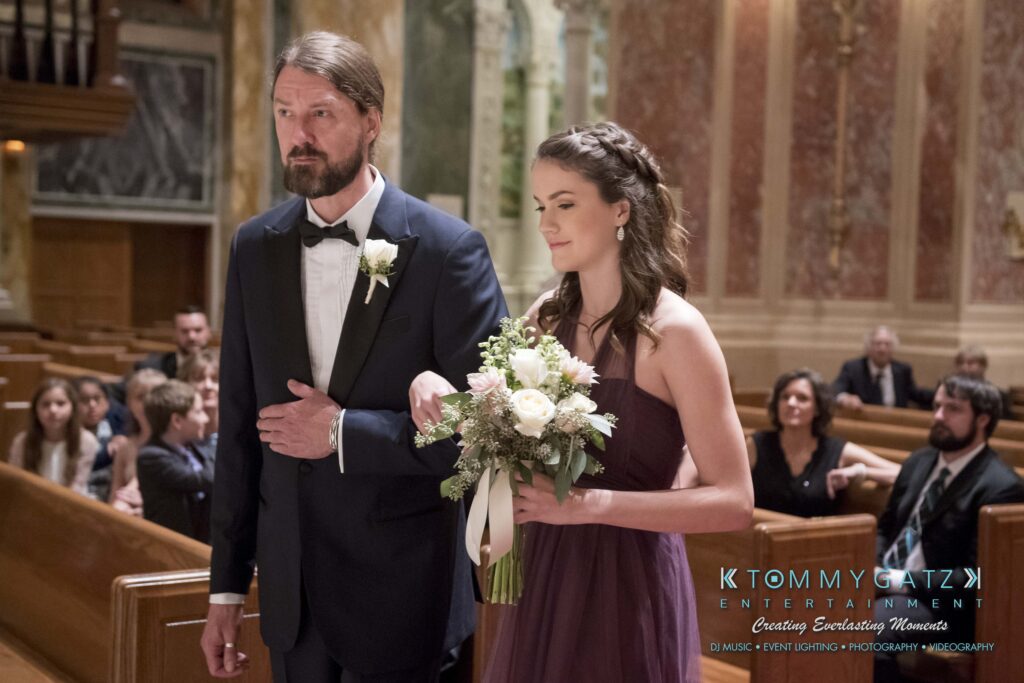 A man in a suit walks down an aisle with a woman in a purple dress holding a bouquet in a church setting.