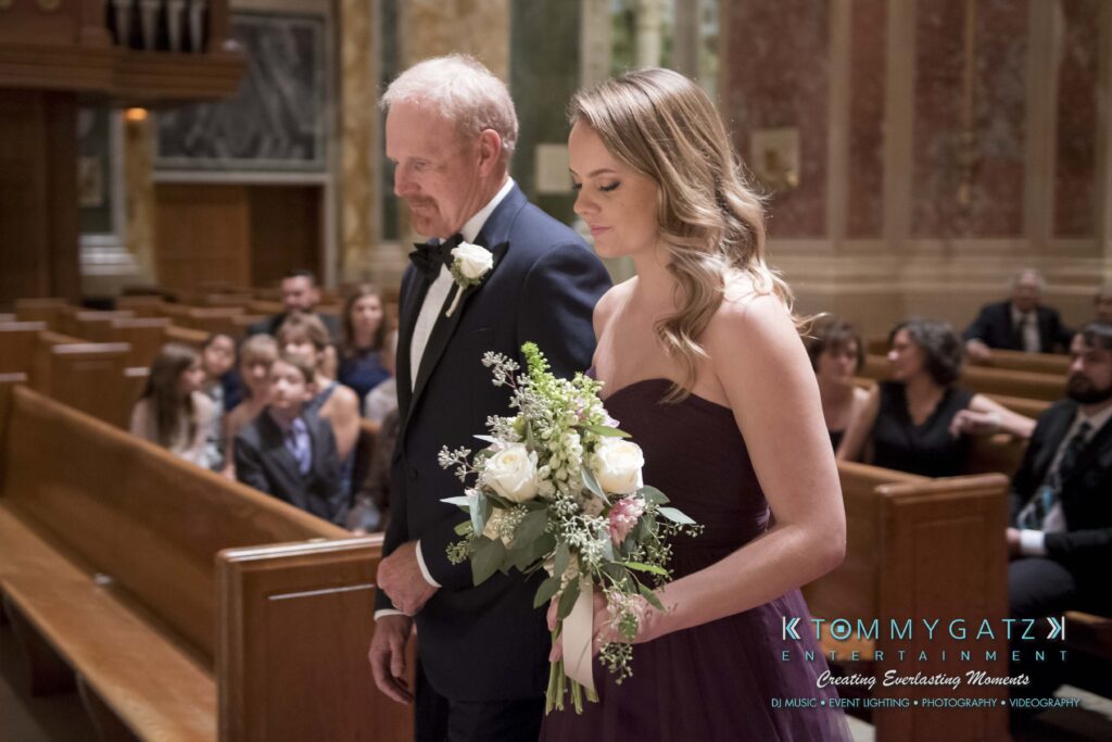 A man and a woman in formal attire walk down a church aisle. The woman holds a bouquet of white roses.