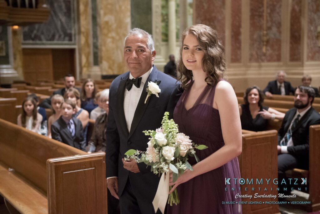 A man in a suit and a woman in a purple dress hold flowers as they walk down the aisle in a church. Guests are seated in pews.