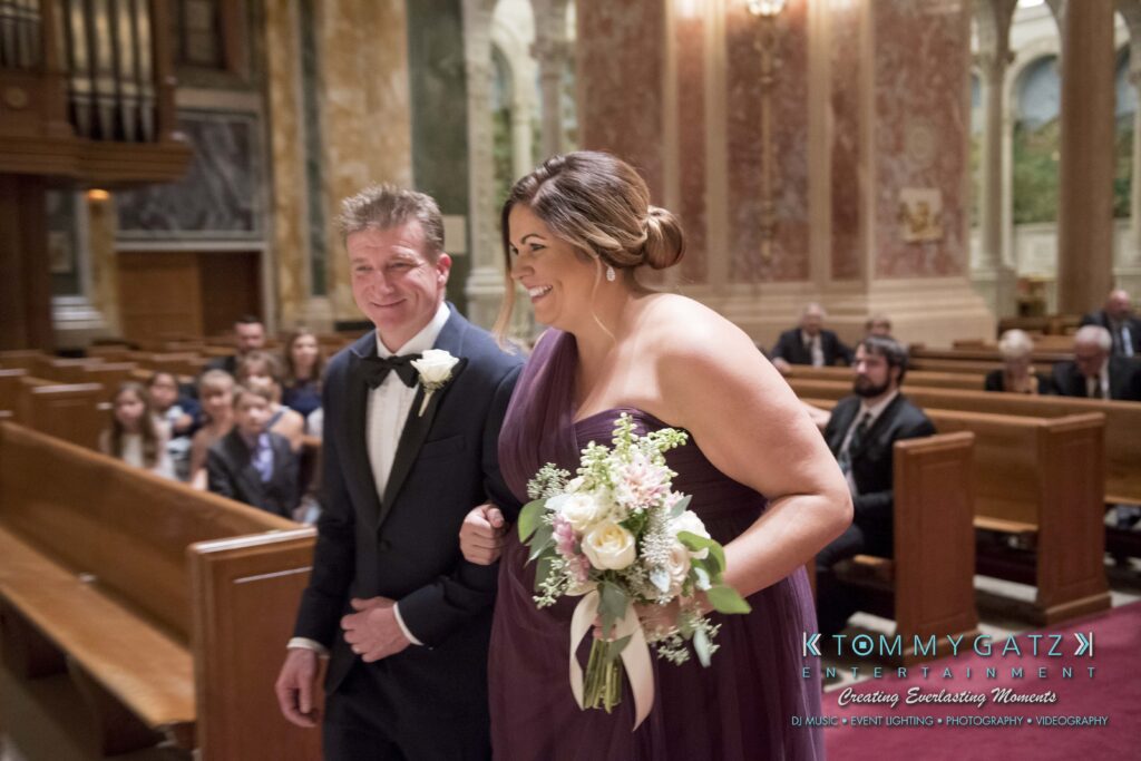 A woman in a purple dress holding a bouquet walks down the aisle with a man in a tuxedo at a formal event in a large ornate venue.