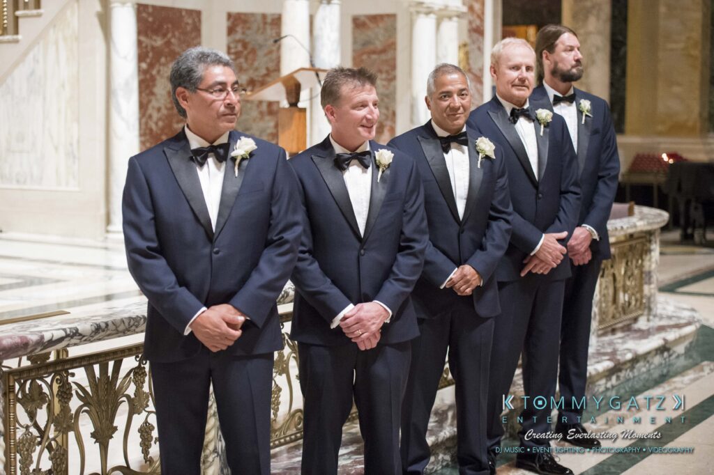 Five men in matching tuxedos stand in a line indoors, smiling, with floral boutonnieres. Marble columns and ornate railing are in the background.