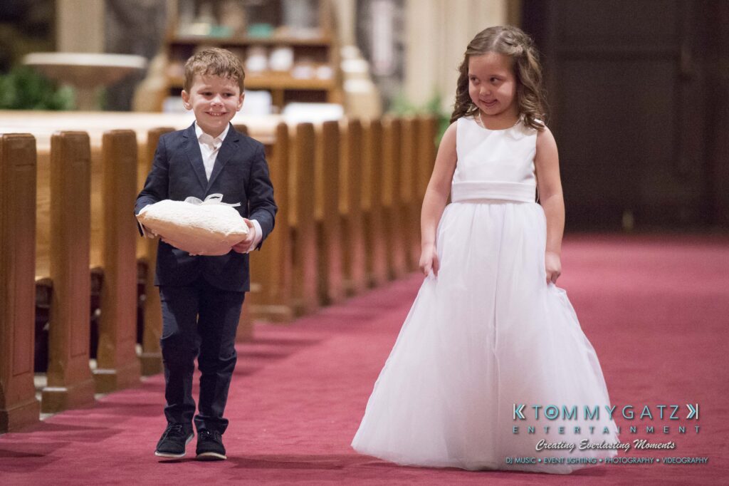 A young boy and girl walk down a church aisle. The boy in a suit holds a pillow, and the girl wears a white dress.