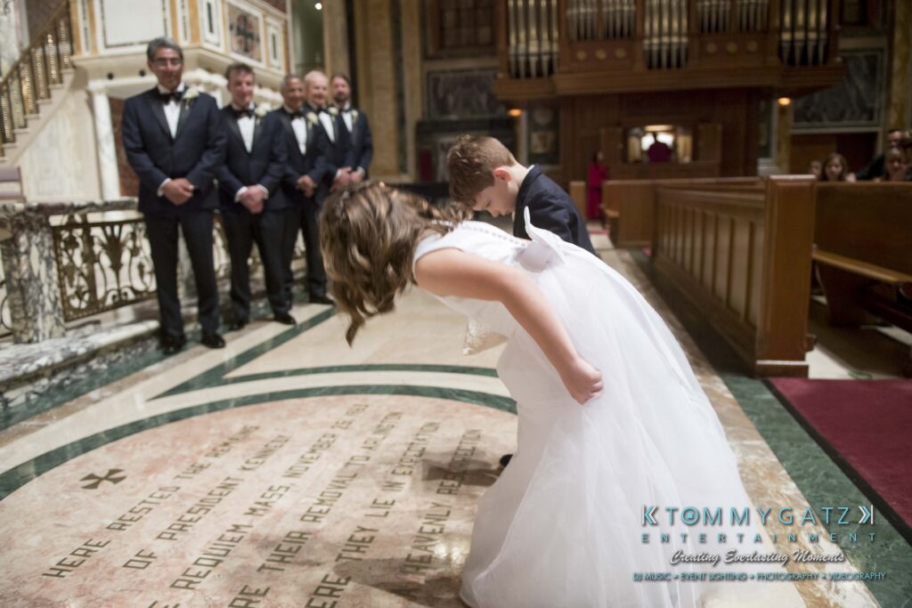 Two children in formal attire bow in front of a church altar during a ceremony, with groomsmen standing in the background.