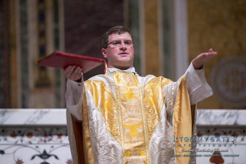 A priest in yellow and white vestments holds an open book while speaking, standing in a church setting with ornate decor.