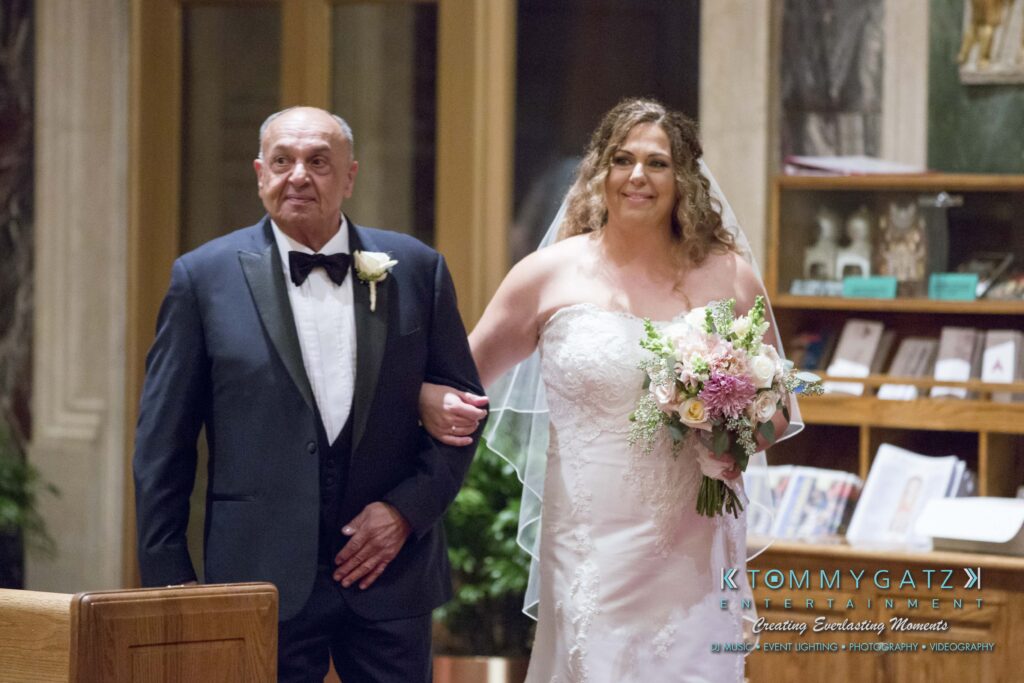 A bride in a white gown and veil holds a bouquet while walking arm-in-arm with an older man in a tuxedo in an indoor setting.