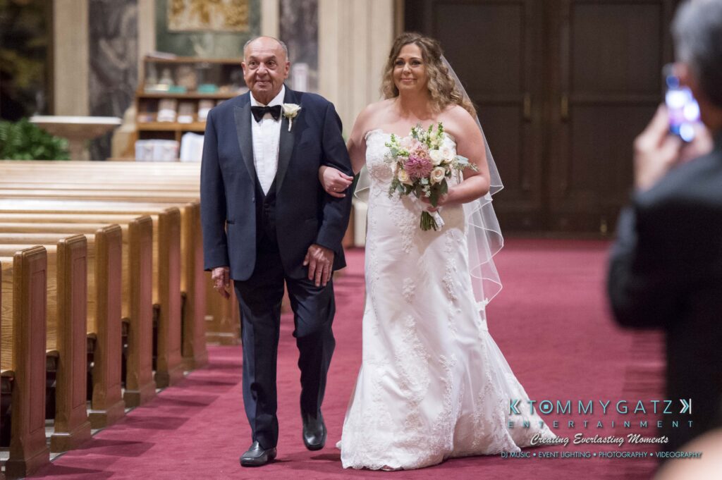 A bride in a white gown walks down the aisle with an older man in a tuxedo inside a church.