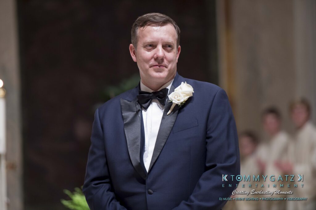 Man in a tuxedo with a white rose boutonniere stands indoors, smiling slightly.